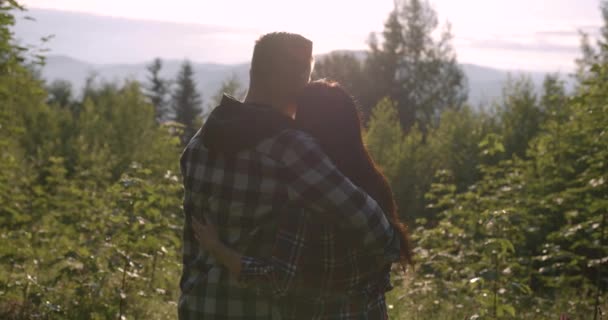 Πλάτη στιγμιότυπο μιας στιγμής ενός ειλικρινούς ρομαντικού ζευγαριού, που υποστηρίζει μαζί, απολαμβάνοντας το ηλιοβασίλεμα, το δάσος και τα βουνά. Μια νεαρή γυναίκα και ένας Καυκάσιος άνδρας σε ένα όμορφο πλεονεκτικό σημείο το καλοκαίρι — Αρχείο Βίντεο
