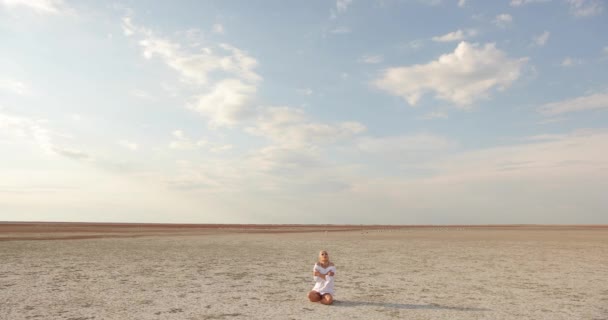 흰 드레스를 입고 하얀 모래 위에 앉아 있는 아름다운 젊은 여성. 사막 상공에 흰 구름이 떠 있는 아름다운 푸른 하늘. — 비디오