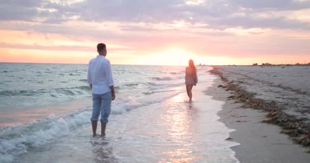 Couple walking at sunset on a summer sea beach. A man in a white shirt stands in the water, a woman in a dress runs along the beach — стоковое видео