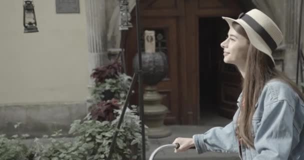 Boční portrét mladé atraktivní brunetky ve stylovém klobouku na kole podél staré ulice zdobené rostlinami. Záběry 4k. — Stock video