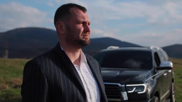 Ein Mann im Businessanzug mit ernstem Gesichtsausdruck steht auf einem Gebirgshintergrund neben einem schwarzen Geländewagen. Geschäftsmann in der Nähe des Autos — Stockvideo