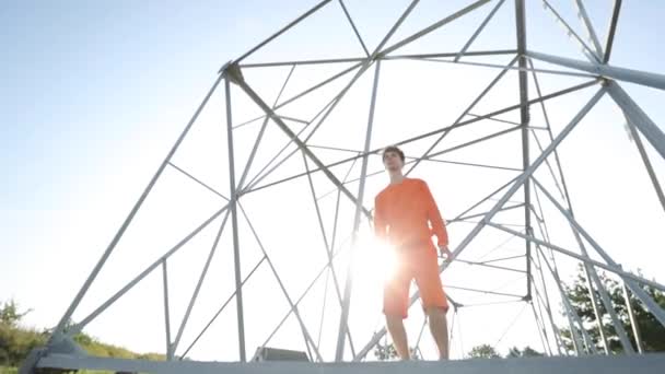 Кудрявый парень в ярко-оранжевом спортивном костюме прыгает рядом с большой металлической структурой современного искусства. Молодой человек в удобной повседневной одежде. — стоковое видео