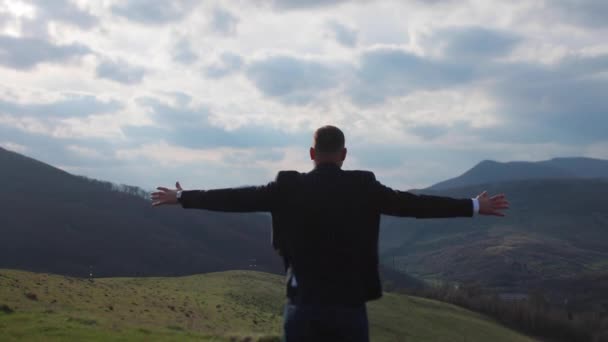 Человек в костюме стоит на фоне прекрасного пейзажа гор, поднимая руки вверх. — стоковое видео