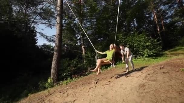 Pasangan muda berayun di bawah pohon hijau pada hari yang cerah di musim semi. Pacar dan pacarnya menikmati musim panas yang romantis. Yong man and woman in love swaying on swing — Stok Video