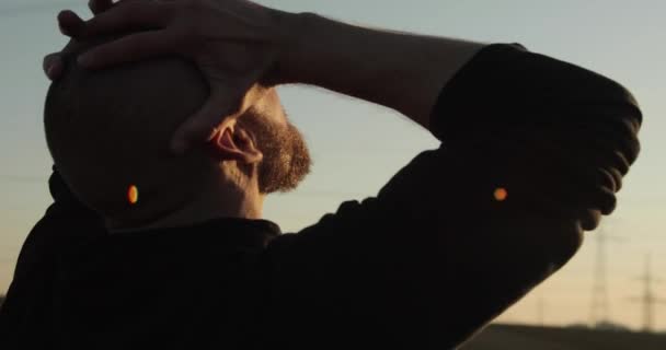 Un joven barbudo calvo permanece entre el campo de trigo al atardecer. Se aferra a su cabeza y la levanta hacia el sol. — Vídeos de Stock