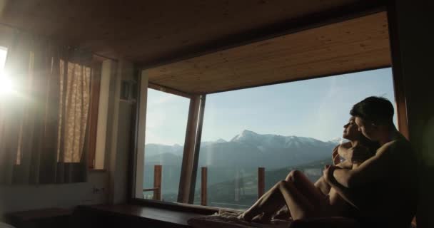 Вид збоку молодої пари без одягу, що обіймається на вікнах, матиме неймовірну панораму на засніжені гори. Дерев'яний котедж в горах . — стокове відео