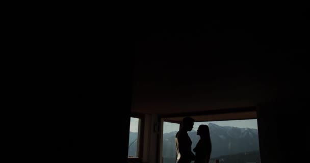 Casal jovem nu ficar perto da janelavontade com panorama incrível em montanhas cobertas de neve e beijando. Casa de madeira nas montanhas. — Vídeo de Stock