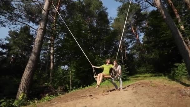 Pasangan muda yang cantik berayun di ayunan di hutan. Muda tersenyum gembira wanita berayun di ayunan di hutan musim panas. — Stok Video