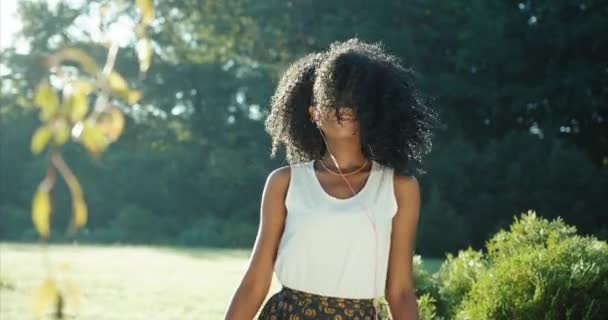 Wesoła, ładna afro-amerykańska dziewczyna z zielonymi cieniami do powiek uśmiecha się, aktywnie tańczy słuchając muzyki w słuchawkach. Lokalizacja ogrodu w słoneczny dzień. — Wideo stockowe