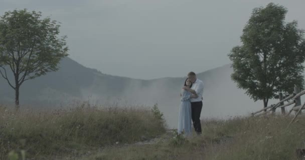 El hombre está abrazando a la hermosa mujer y acariciando su mejilla mientras está de pie en el camino del prado en las montañas cubiertas de espesa niebla. — Vídeo de stock