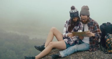 Dağlara git. Büyüleyici genç hipster çift kalın bir sis ile kaplı kayanın üzerinde oturur ve bir harita öğrenir.