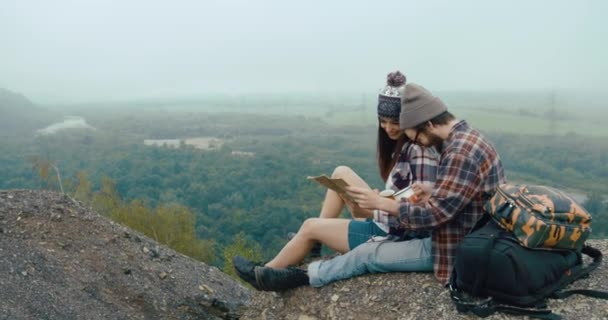 到山上去旅行。迷人的年轻催眠夫妇坐在布满浓雾的岩石上学习地图 — 图库视频影像