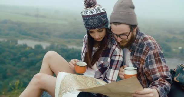 Reis opp i fjellene. Sjarmerende, unge hypsterpar sitter på en klippe dekket av tykk tåke, drikker varm kaffe og lærer et kart. – stockvideo