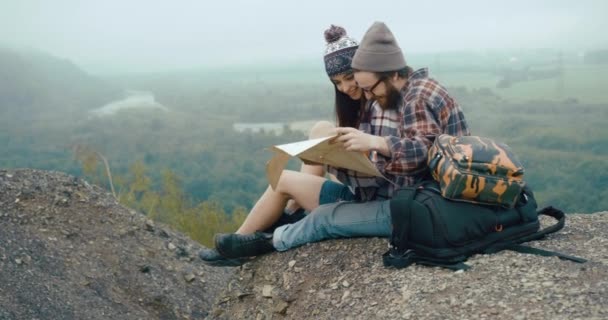 Viaje para as montanhas. Encantador jovem casal hipster senta-se na rocha coberta com névoa espessa, bebe café quente e aprende um mapa — Vídeo de Stock