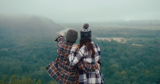 Călătoreşte în munţi. Bărbat și femeie îmbrăcați în stil hipster se uită la priveliștea montană de pe stânca acoperită cu ceață și se îmbrățișează reciproc tandru — Videoclip de stoc