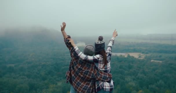 Perjalanan ke pegunungan. Pria dan wanita berpakaian hypster melihat pemandangan gunung dari batu tertutup kabut dan mengangkat tangan mereka ke atas loke pemenang — Stok Video