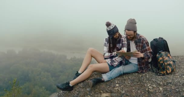 Matkusta vuorille. Viehättävä nuori hypsteripari istuu kalliolla paksun sumun peitossa ja oppii kartan. — kuvapankkivideo