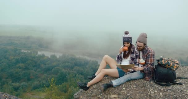 Reis opp i fjellene. Sjarmerende, unge hypsterpar sitter på en klippe dekket av tykk tåke og lærer et kart. – stockvideo