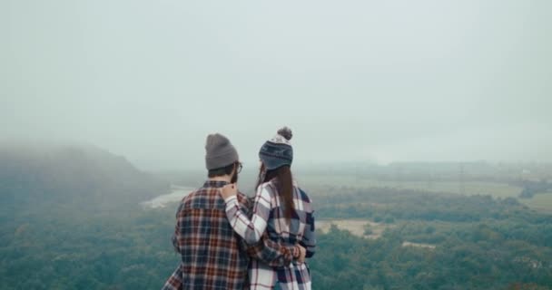 Viaja a las montañas. Hombre y mujer vestidos con estilo hipster miran la vista de la montaña desde la roca cubierta de niebla y se abrazan tiernos — Vídeo de stock