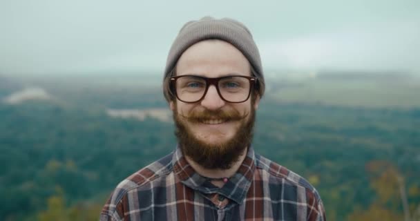 Uśmiechnięty młody człowiek z wąsami i brodą stoi na szczycie skały z pięknym górskim krajobrazem za — Wideo stockowe