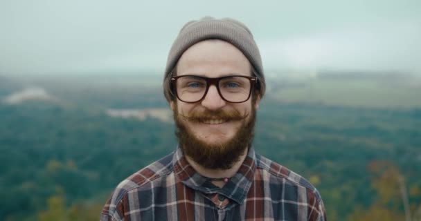 Tersenyum pemuda dengan kumis dan jenggot berdiri di atas batu dengan pemandangan gunung yang indah di belakang — Stok Video