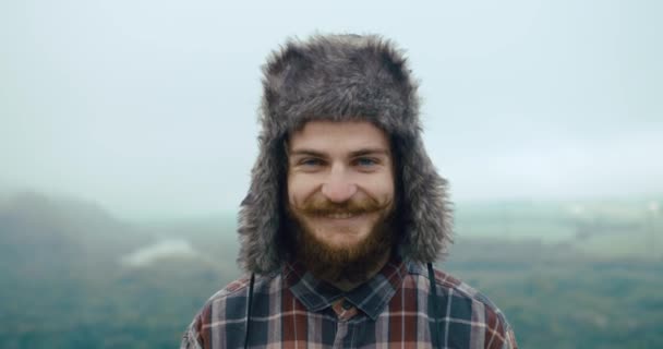 Jovem sorridente com bigodes e barba fica no topo de uma rocha com bela paisagem montanhosa atrás — Vídeo de Stock