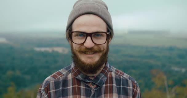 Uśmiechnięty młody człowiek z wąsami i brodą stoi na szczycie skały z pięknym górskim krajobrazem za — Wideo stockowe