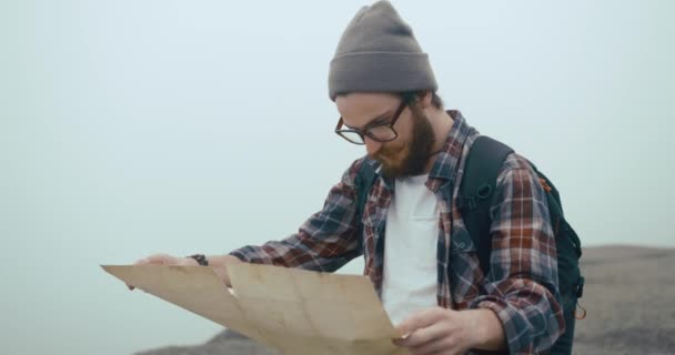 Unterwegs in den Bergen. Junger Mann mit stylischem Bart lernt alte Landkarte beim Wandern auf dem mit dichtem Nebel bedeckten Berg — Stockvideo