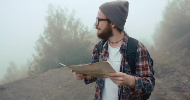 Viajando por las montañas. Joven con barba elegante aprende un viejo mapa caminando por la montaña cubierto de espesa niebla — Vídeos de Stock