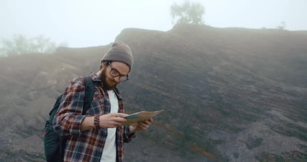 Reiser rundt i fjellene. Unge mann med stilig skjegg lærer et gammelt kart som går på fjellet dekket av tykk tåke – stockvideo