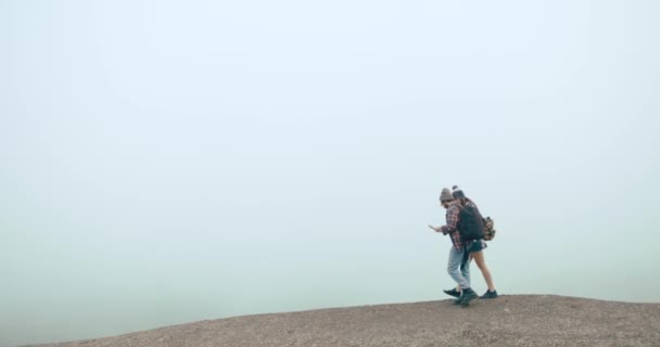 Reis opp i fjellene. Gutt og jente kledd som hypstere går med kart rundt fjellene dekket av tykk tåke – stockvideo