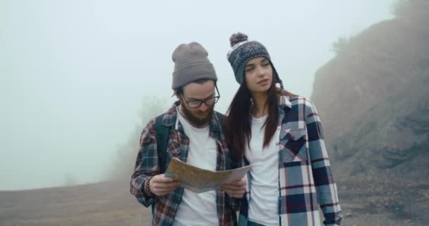 Matkusta vuorille. Poika ja tyttö pukeutunut hypsterit kävellä kartta ympäri vuoria peitetty paksu sumu — kuvapankkivideo