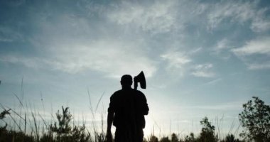 Omzunda baltası olan bir oduncunun silueti kameradan uzağa güneşe doğru hareket ediyor.