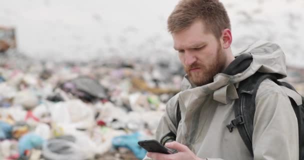 Οικολογική καταστροφή. Ένας νεαρός γενειοφόρος στέκεται σε μια χωματερή και χρησιμοποιεί ένα smartphone. οικολογική καταστροφή. ρύπανση. — Αρχείο Βίντεο