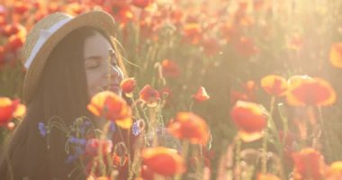 Gün batımında haşhaş tarlasının ortasında gülümseyen kadın. Hasır şapkalı esmer bayan, haşhaş kokluyor. Yaz günü ve sakin bir atmosfer.