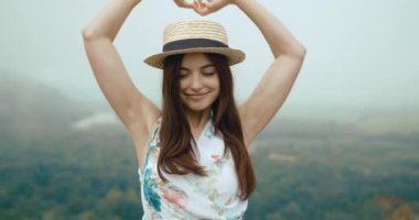 Yazlık şapkası ve elbisesi olan alımlı genç bir kadın arkasında muhteşem bir manzarayla bir dağın tepesinde duruyor..