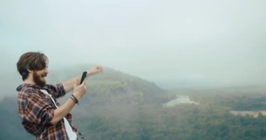 Genç hipster adam, güzel dağ manzarası sisle kaplanmadan önce telefonuyla kayanın tepesinde duran biriyle sohbet eder ve kazanan gibi elini kaldırır..