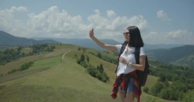 Young smiling brunette tourist with natural make-up and backpack is taking selfie via the mobile phone while hiking in green mountains in summer. 4k footage