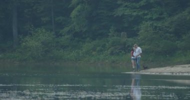 The beautiful happy loving couple are tenderly hugging and rubbing noses while standing with their dog on the riverbank at the background of the forest. The man is kissing his lover in head