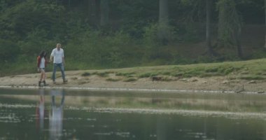 Romantic walk of the happy beautiful loving couple holding hands and talking while walking with their dog along the river bank near the forest. 4k footage