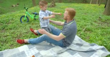 Beautiful family portrait of the pretty cheerful son running to father and hugging him during the picnic in forest. They are falling down on the planket. 4k footage