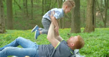 Happy father is raising up and spinning round his smiling little son while lying on the blanket during the picnic in forest