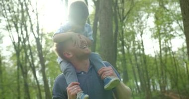 The happy smiling father is giving the piggyback ride to his little son. The cute boy is closing father eyes. Forest location. 4k footage