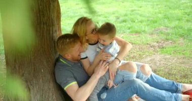 Family portrait. Happy parents are hugging and tickling their little smiling son while sitting under the tree on the grass. 4k footage