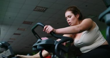 The exhausted obese woman is hardly working out on the exercise bike and sadly leaning on her arms. Gym location. 4k.