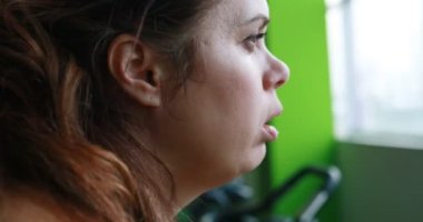 Close-up view of the tired overweight woman activily working out on the exercise bike in the green gym. Moving footage down