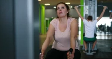 Portrait of the obese woman actively working out on the rowing machine in the green gym. Camera is moving left and right