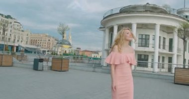 Attractive happy woman with blonde hair and natural make-up in pink dress is enjoying the weather. She is spinning round while walking along the sunny street
