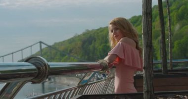 Portrait of the attractive charming blonde girl standing on the balcony and enjoying the view of the beautiful river and mountains covered with forests