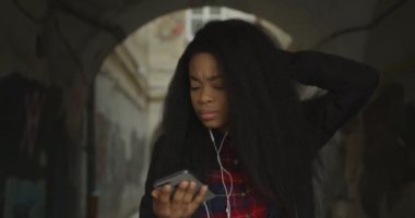 Beautiful african girl with curly hair is having fun outdoor. She is enjoying music in earphones and actively dancing. 4k footage